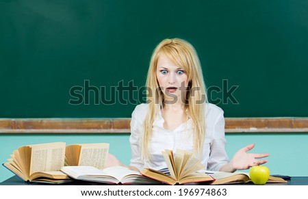 Closeup portrait hopeless skeptical overwhelmed student sitting at desk in front pile book studying preparing for finals isolated chalkboard background. Education college life concept. Face expression