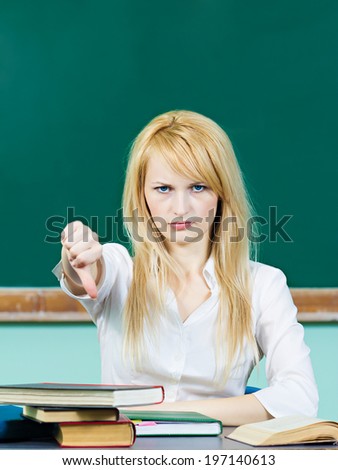 Closeup portrait angry pissed off woman, annoyed student, sitting at desk in classroom giving thumbs down looking unhappy, negative face expression, disapproval isolated chalkboard background. Emotion