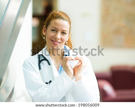 Closeup portrait, beautiful smiling cheerful health care professional, pharmacist, dentist, nurse making heart sign hands isolated background hospital hallway. Positive human emotion facial expression