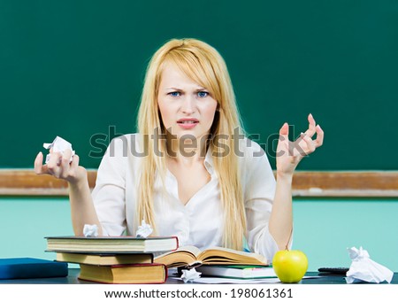 Closeup portrait hopeless skeptical overwhelmed student sitting at desk book, crumpled paper, studying preparing finals isolated chalkboard background. Education college life concept. Face expression