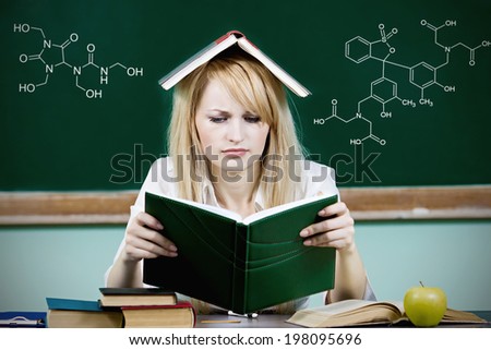 Closeup portrait hopeless overwhelmed student sitting at desk, reading book studying preparing finals isolated chalkboard background with chemistry formulas. Education college concept. Face expression