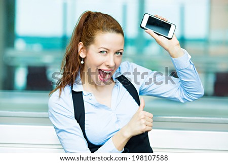 Closeup portrait happy, excited, young, attractive business woman, company employee showing, holding her smart phone, giving thumbs up, isolated background windows corporate office, building. Emotions