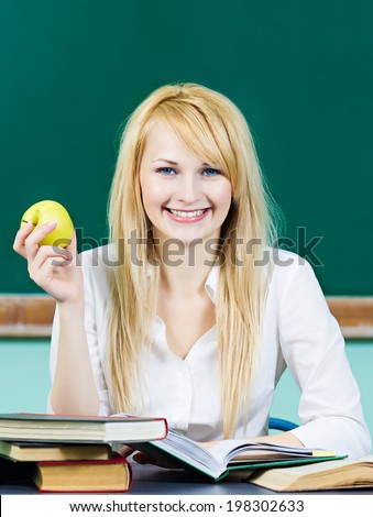 Closeup portrait attractive young happy smiling woman, student sitting at desk with text books, in classroom holding apple isolated chalkboard background. Education college concept. Positive emotion
