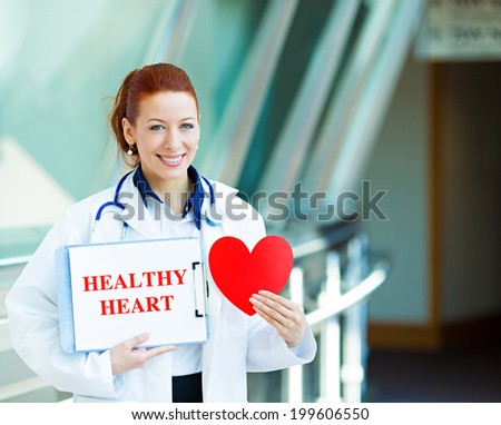 Closeup portrait happy smiling female health care professional, woman family doctor, cardiologist with stethoscope holding sign healthy heart, heart isolated hospital hallway background. Patient plan