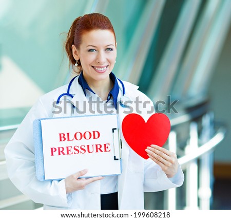 Closeup portrait happy smiling female health care professional, woman family doctor, cardiologist with stethoscope holding sign blood pressure, heart isolated hospital hallway background. Patient plan