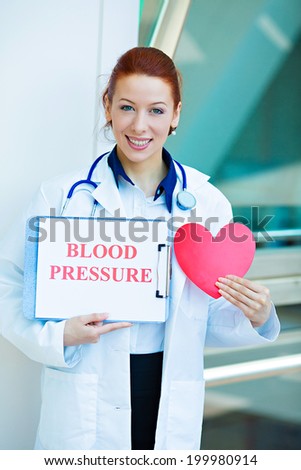 Closeup portrait happy smiling female health care professional, woman family doctor, cardiologist with stethoscope holding heart, blood pressure sign isolated hospital hallway background. Patient plan
