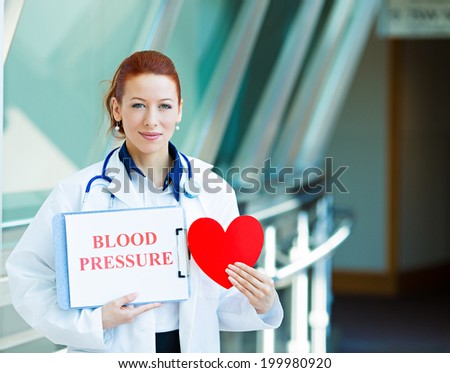 Closeup portrait happy smiling female health care professional, woman family doctor, cardiologist with stethoscope holding heart, blood pressure sign isolated hospital hallway background. Patient plan