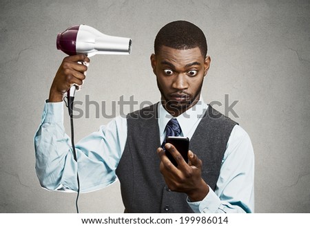 Closeup portrait worried, shocked business man, deal maker reading bad news on smart, mobile phone holding hairdryer isolated black grey background. Human face expression, emotion, corporate executive