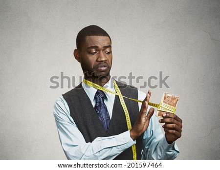Closeup portrait sad, young, confused man holding, looking at fatty, sweet cookie with measuring tape around, trying to withstand resist temptation to eat it isolated black background. Face expression