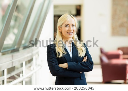 Closeup portrait, young beautiful blonde business woman in black suit smiling isolated on corporate office hallway background. Positive human emotions, facial expressions, attitude, life perception