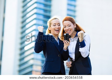 Portrait happy business women, arms raised up, partners receiving news on mobile phone, looking at smartphone reading text message pumping fist celebrating success
