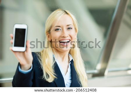 Closeup portrait happy, excited, young, attractive business woman, company employee showing, holding her smart phone isolated background windows corporate office. Positive emotion reaction, perception