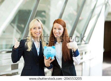 Closeup portrait two smiling business women bank employees holding piggy bank giving thumbs up isolated corporate office background. Financial savings banking concept. Positive emotion face expression