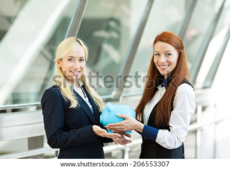 Closeup portrait two happy smiling business women, bank employee holding giving piggy bank to customer isolated corporate office background. Financial savings concept. Positive emotion face expression