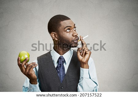 Closeup portrait headshot corporate executive businessman making bad health choices, smoking cigarette instead of having fresh green apple isolated black background. Face expressions, body language