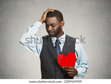 Closeup portrait young sad confused corporate man, holding red heart in his hands, about to cry, looking up isolated grey, black background. Negative human emotion facial expression feelings, attitude