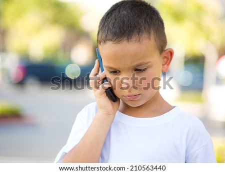 Closeup portrait, worried, sad young child, boy talking on phone to someone, looking unhappy, isolated outdoors background. Negative human emotions, facial expressions, feeling, reaction Communication