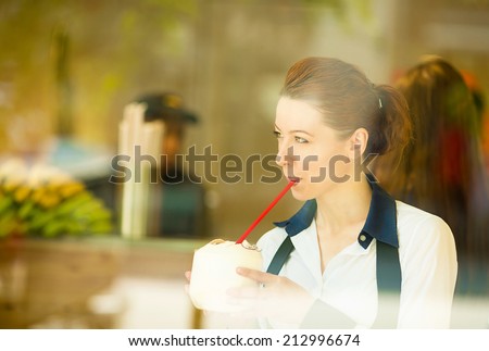 Cafe city lifestyle woman drinking coconut juice, looking outside on street through glass window, sitting indoor juice bar. Cool young modern female model in her 20s. Positive face expression, emotion