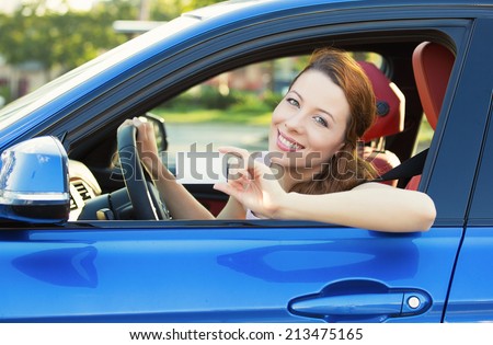 Young woman in new car showing blank drivers license or sign out, through side car window. Happy lovely female model driver enjoying her new auto outside parking lot. Positive face expression, emotion