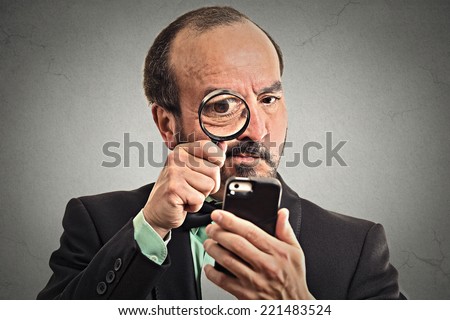 Curious. Mature business man looking through a magnifying glass on smart phone isolated grey wall background. Human face expression. Investigator looking with magnifying glass. Security safety concept