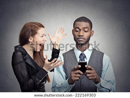 Upset angry woman trying to bring attention of young handsome man ignoring her looking at smartphone reading browsing internet isolated grey wall background. Phone addiction concept. face expression