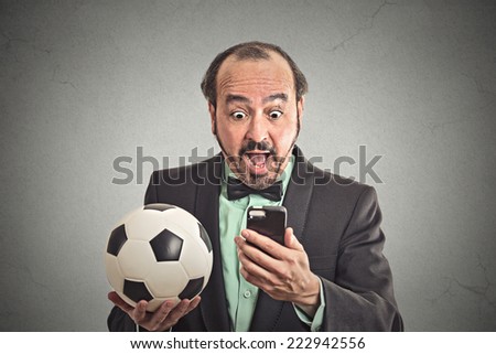 Portrait excited man looking shocked with opened mouth on cell smart phone watching  game holding football isolated grey wall background. Positive emotion, face expression. Data plan online gaming