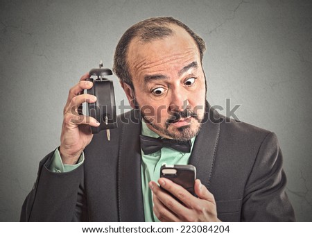 surprised man with alarm clock looking at his smart phone with confused face expression isolated grey wall background