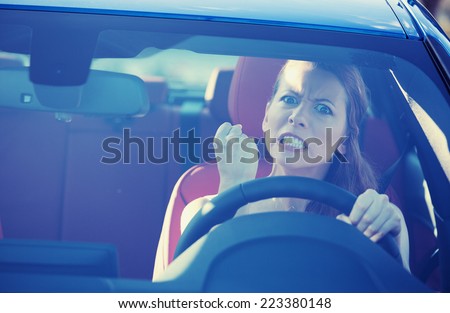 Portrait displeased angry pissed off aggressive woman driving car, shouting at someone in traffic hand fist up in air front windshield view. Emotional intelligence concept. Negative human expression