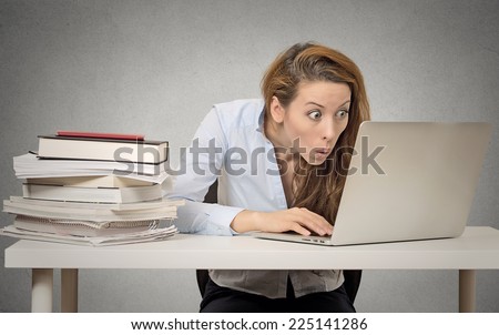 Woman using her laptop computer sitting at table with pile of books on grey wall office background. New technology internet addiction mania concept. Funny looking girl obsessed with pc. Human emotions