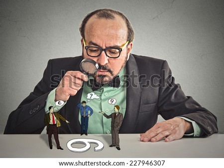 Businessman with glasses sitting at desk skeptically looking at arguing people through magnifying glass isolated grey office wall background. Human face expression, attitude. To each its own concept