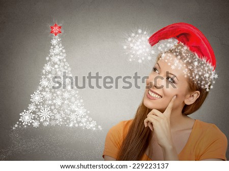 x-mas, winter, happiness concept smiling woman in red santa helper hat looking at christmas tree on grey wall background. Positive emotion face expression perception vision