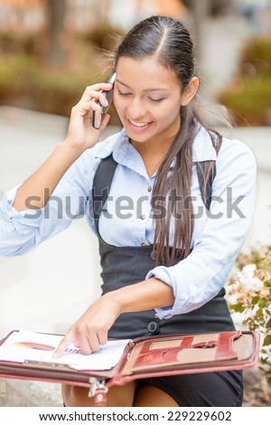 Closeup portrait beautiful smiling young businesswoman in blue shirt sitting outside reviewing financial statements paperwork talking on mobile phone isolated park trees background. Facial expression