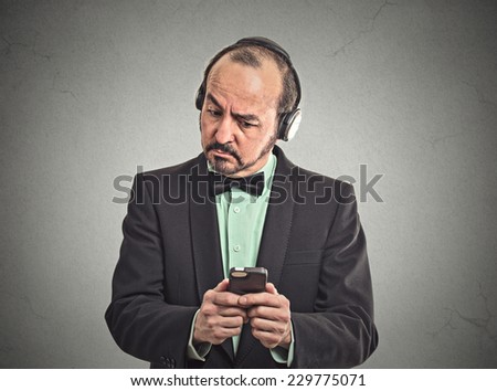 Confused middle aged man listening music with pair of headphones looking at his smartphone surprised. Closeup portrait man reading news on mobile phone isolated grey wall background. Face expression
