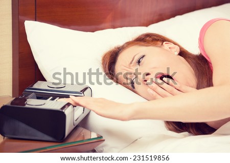 Closeup portrait woman extending hand to alarm clock yawning lies in bed trying to wake up for new day. Human face expression, emotion, feeling. Sleep deprivation, lack of adequate sleep concept