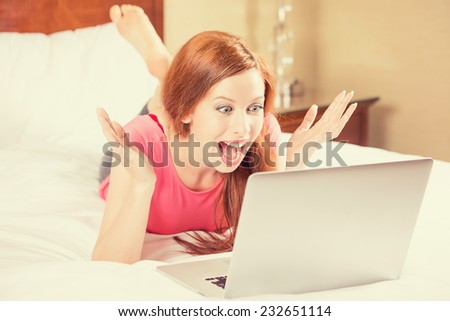 Excited young beautiful woman with arms raised using looking at her laptop screen laying in bed. Positive human emotion happy facial expression, body language, reaction. Unexpected pleasant  good news