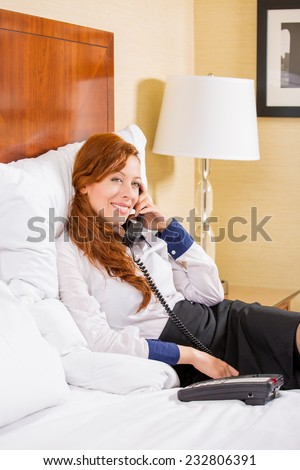 Taking time to talk with nearest. Beautiful young smiling businesswoman in white shirt and dress talking on phone while sitting on the bed in hotel room. Travel, communication, socializing concept
