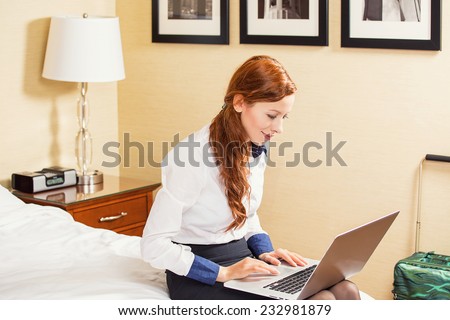 Staying in touch with my colleagues. Top view of beautiful young businesswoman in skirt and white shirt working on laptop and smiling while sitting on the bed in hotel room. Positive face expression