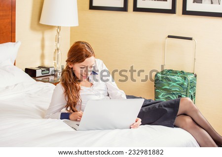 Staying in touch with my colleagues. Top view of beautiful young businesswoman in skirt and white shirt working on laptop and smiling while sitting on the bed in hotel room. Positive face expression