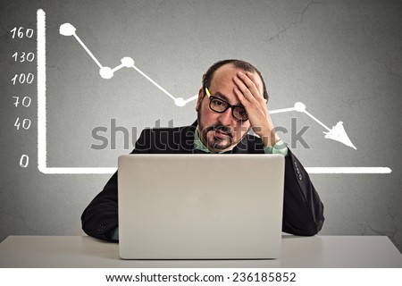 Frustrated stressed business man sitting at table in front of computer with financial market chart graphic going down on grey office wall background. Poor economy concept. Face expression, emotion