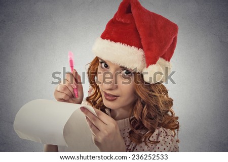 Portrait beautiful woman with christmas hat is posing in studio writing gift ideas on roll of paper isolated on grey wall background. holiday season, xmas shopping ideas. Positive face expression