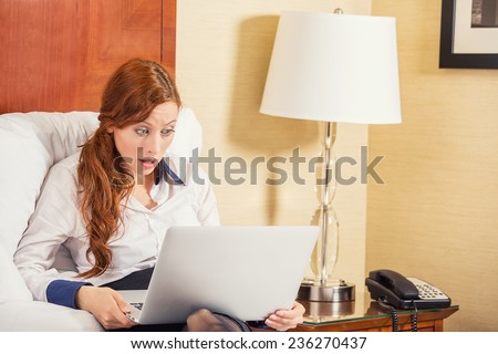 Shocked young business woman using laptop looking at computer screen blown away in stupor sitting in bed at home, hotel. Human face expression, emotion, feeling, perception, body language, reaction