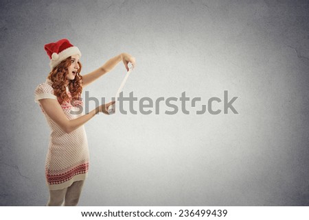 Portrait young attractive girl santa helper in red hat holding reading christmas wish list isolated grey wall background with copy space. Holiday season celebration tradition concept. Positive emotion