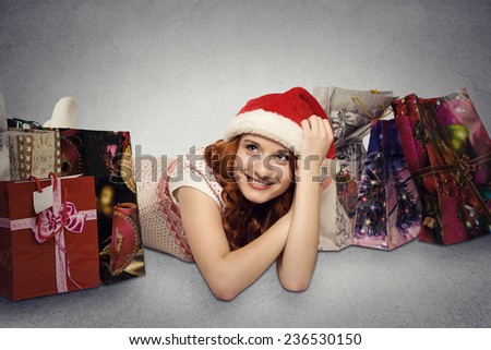 Happy woman in red santa claus hat with christmas gifts dreaming fun holidays. Young girl looking up smiling with shopping bags isolated grey wall background with copy space. Positive face expression