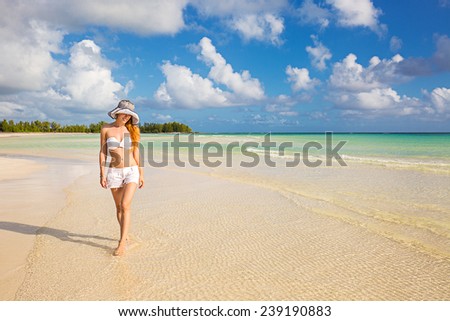 Young happy smiling woman in white shirts and hat walking on beach enjoying tropical weather ocean view. Holiday vacation getaway concept. Healthy life style well being.