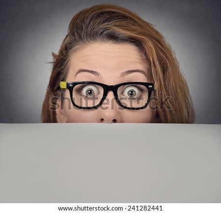 Banner sign woman peeking over edge of blank empty paper billboard. Beautiful young woman with glasses looking surprised and scared funny with wide open eyes isolated on grey wall background
