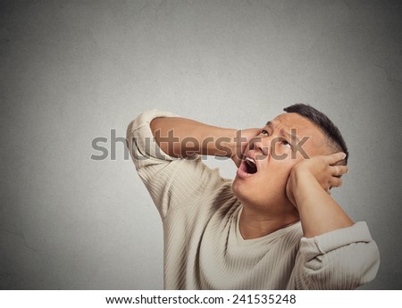 Closeup portrait angry mad unhappy stressed man covering his ears looking up, stop making loud noise it's giving me headache isolated grey wall background. Negative human  emotion, face expression
