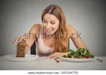Portrait young woman deciding whether to eat healthy food or sweet cookies she is craving sitting at table isolated grey wall background. Human face expression emotion reaction Diet nutrition concept