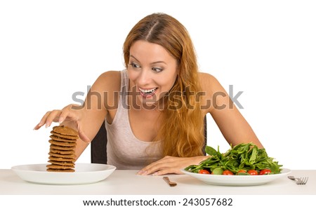 Portrait young woman deciding whether to eat healthy food or sweet cookies she is craving sitting at table isolated white background. Human face expression emotion reaction. Diet nutrition concept