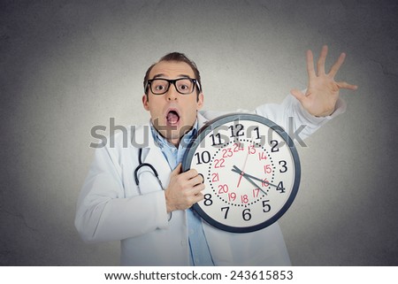 Closeup portrait overwhelmed, busy, unhappy male health care professional, funny looking doctor guy holding wall clock running out of time exhausted isolated grey wall background. Healthcare reform