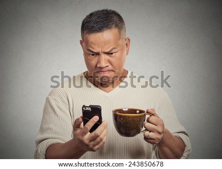 Closeup portrait worried angry frustrated man reading bad news sms on smart mobile phone drinking holding cup coffee tea isolated grey wall background. Human face expression emotion reaction